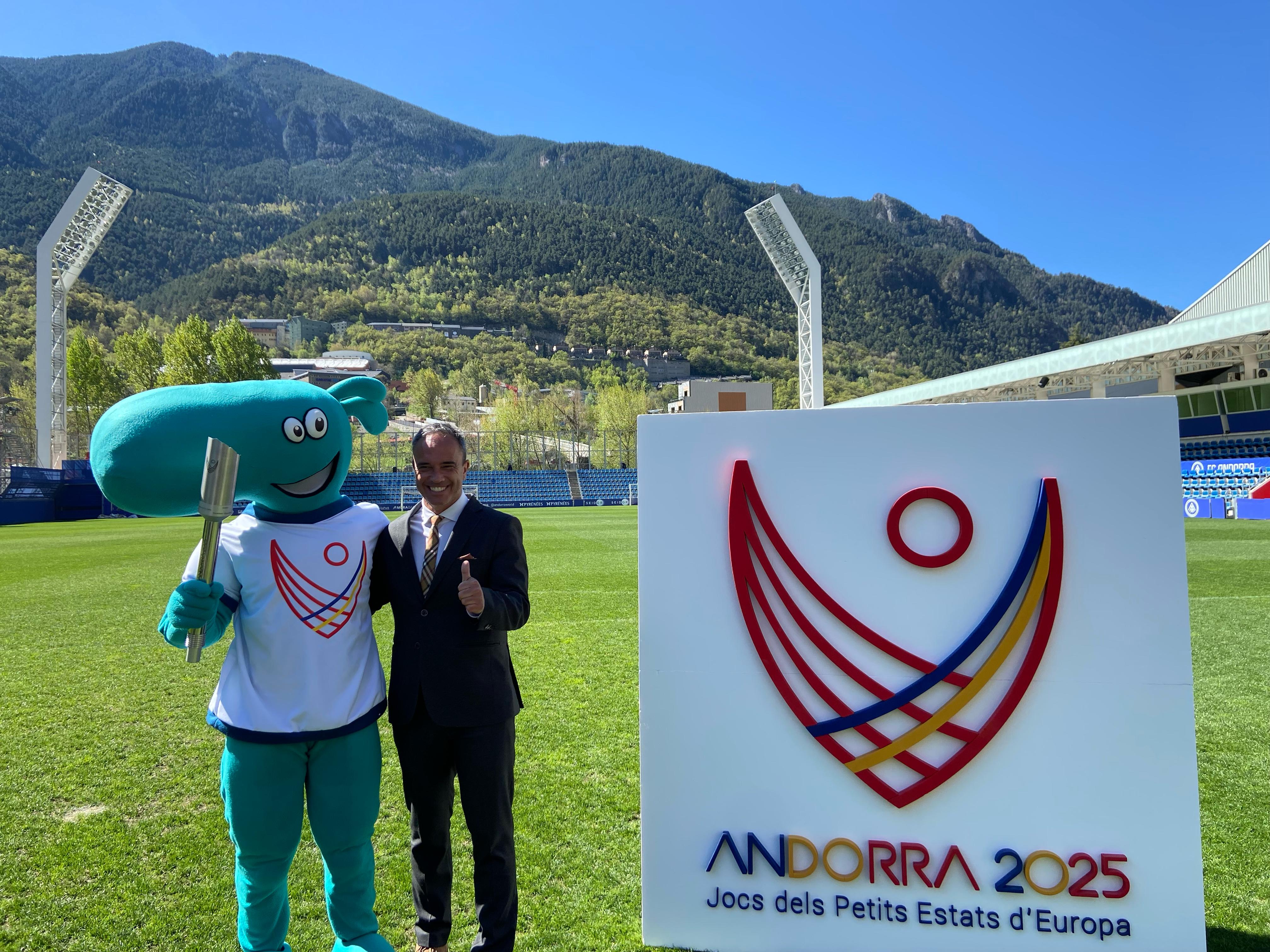 El secretari d'Estat, Alain Cabanes, amb la mascota, la torxa i el logotip d'Andorra 2025. El secretari d'Estat, Alain Cabanes, amb la mascota, la torxa i el logotip d'Andorra 2025.